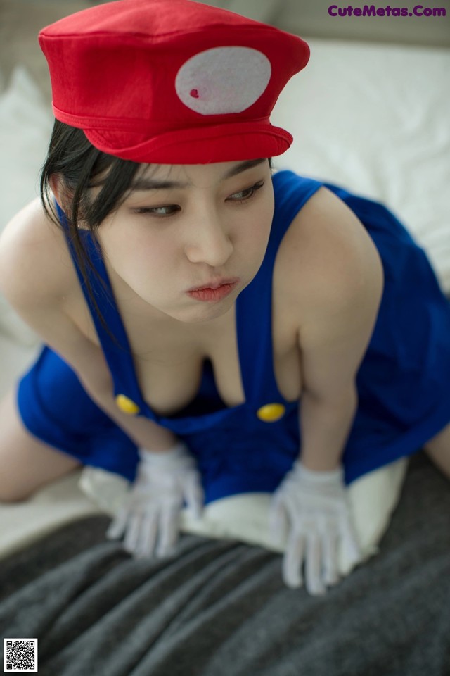 A woman in a red hat and blue overalls sitting on a bed.